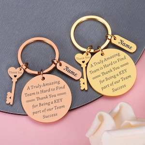 May include: Two metal keyrings, one rose gold and one gold, each featuring a round disc engraved with 'A Truly Amazing Team is Hard to Find Thank You for Being a KEY part of our Team Success', a heart-shaped key charm, and a rectangular name tag.