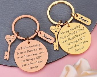 May include: Two metal keyrings, one rose gold and one gold, each featuring a round disc engraved with 'A Truly Amazing Team is Hard to Find Thank You for Being a KEY part of our Team Success', a heart-shaped key charm, and a rectangular name tag.