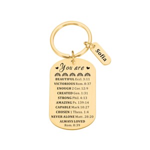 Puede incluir: Llavero dorado con una etiqueta en forma de coraz&oacute;n que dice "You are" seguido de una lista de afirmaciones positivas y referencias b&iacute;blicas. La etiqueta est&aacute; unida a un anillo dorado y a una etiqueta m&aacute;s peque&ntilde;a con el nombre "Sofia" en ella.