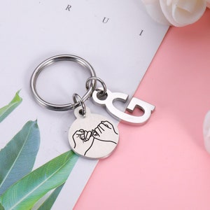 May include: Silver keychain with a small charm featuring two hands making a pinky promise. The charm is attached to a silver ring and a larger charm with the letter "G" on it.
