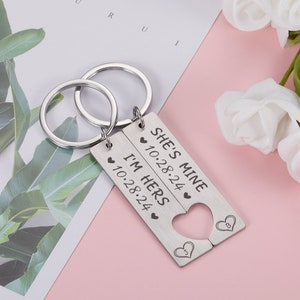 May include: A set of two silver keychains with interlocking heart-shaped charms. The charms are engraved with the text "She's Mine 10.28.24" and "I'm Hers 10.28.24".