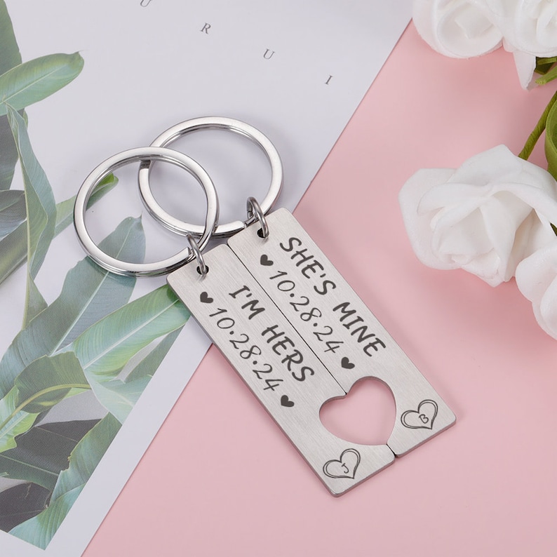 May include: A set of two silver keychains with interlocking heart-shaped charms. The charms are engraved with the text "She's Mine 10.28.24" and "I'm Hers 10.28.24".