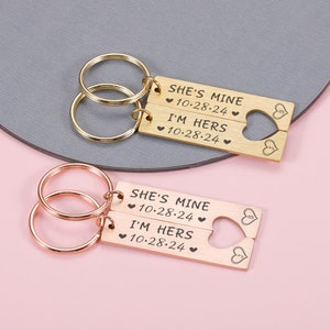 May include: Two gold-tone keychains with interlocking heart-shaped charms. The charms are engraved with the text "She's Mine 10-28-24" and "I'm Hers 10-28-24".