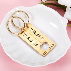 May include: Gold-toned keychain with two interlocking rings and a split heart charm. The charm has the date "12.08.18" engraved on each side and the initials "B&G" on the bottom.