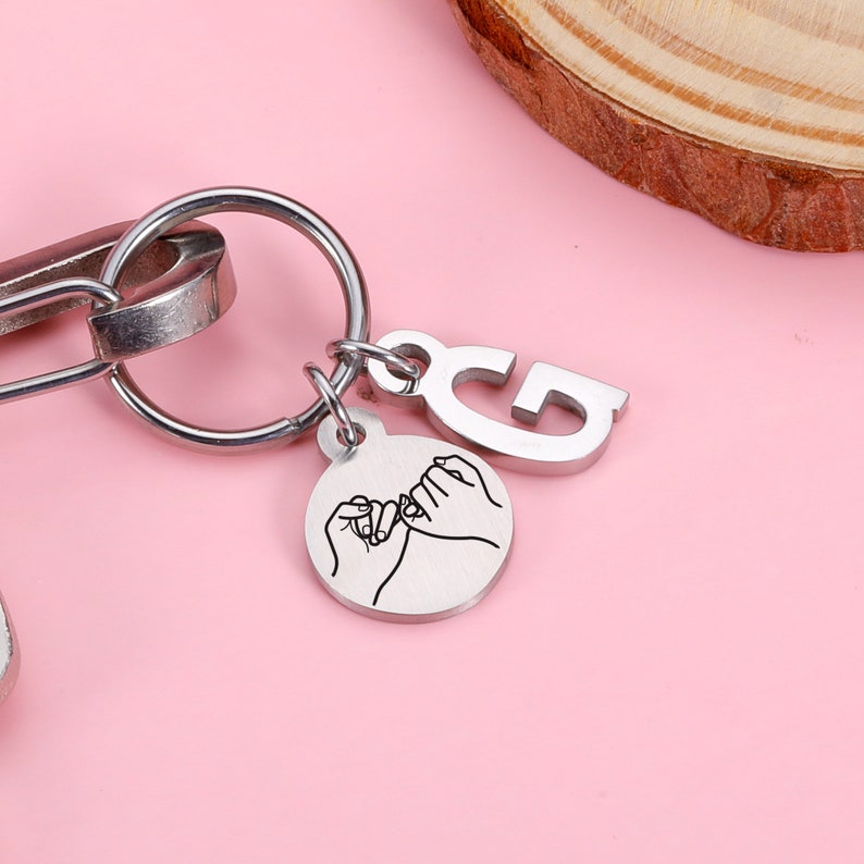 May include: Silver keychain with a small round charm featuring two hands intertwined with a pinky promise. The charm is attached to a silver keyring with a letter 'G' charm.