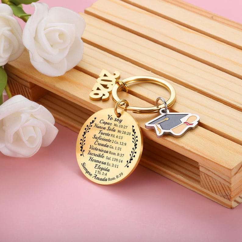 May include: A gold-toned circular keychain featuring the text 'Yo soy' followed by a list of Spanish phrases and Bible verses, a '2026' charm, and a small silver-toned graduation cap charm with a diploma, all attached to a gold-toned metal ring.