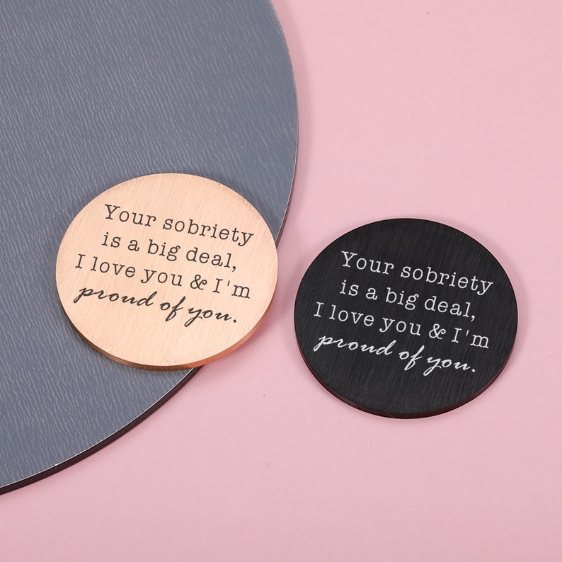 May include: Two round wooden coasters with the text "Your sobriety is a big deal, I love you & I'm proud of you." One coaster is black and the other is copper.
