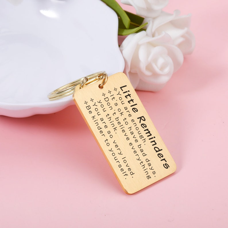 Op de afbeelding: Gouden sleutelhanger met een rechthoekig label met de tekst "Kleine herinneringen" en de tekst "Je bent genoeg. Het is ok&eacute; om slechte dagen te hebben. Geloof niet alles wat je denkt. Je bent zo geliefd. Wees vriendelijker tegen jezelf."