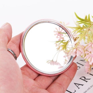 May include: A small, round, pink-rimmed mirror with a silver frame. The mirror is held in a hand.