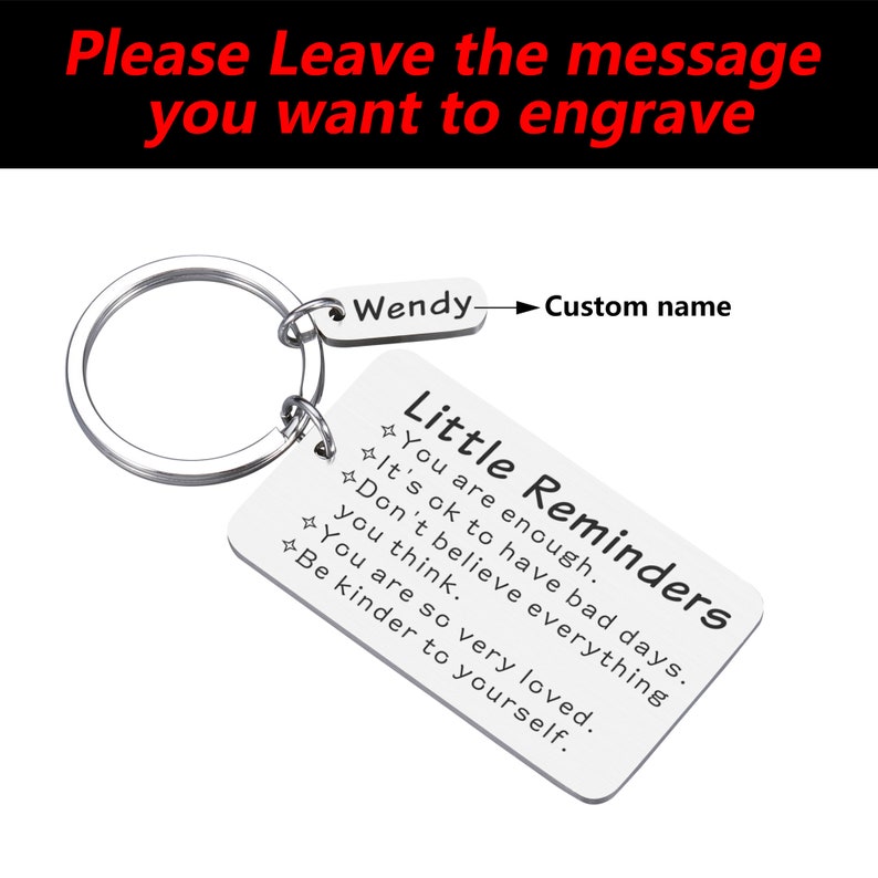 Op de afbeelding: Een zilveren sleutelhanger met een klein rechthoekig label waarop "Little Reminders" staat en de tekst "You are enough. It's ok to have bad days. Don't believe everything you think. You are so very loved. Be kinder to yourself." De sleutelhanger heeft ook een kleiner label waarop "Wendy" staat en een pijl naar rechts die "Custom name" zegt.
