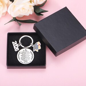 May include: A silver-toned metal keychain featuring a circular pendant engraved with Spanish text and Bible verses, a 2026 charm, and a graduation cap charm. The keychain is presented inside a black square gift box with a black foam insert.