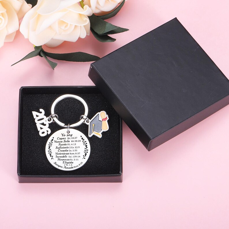 May include: A silver-toned metal keychain featuring a circular pendant engraved with Spanish text and Bible verses, a 2026 charm, and a graduation cap charm. The keychain is presented inside a black square gift box with a black foam insert.