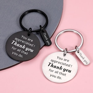 Op de afbeelding: Twee sleutelhangers met ronde metalen labels waarop staat "You are appreciated! Thank you for all that you do." De ene sleutelhanger is zwart en de andere is zilver.