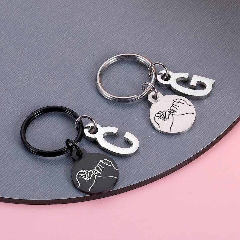 May include: Two silver keychains with black and silver rings. Each keychain has a silver charm with the letter "C" and "G" and a round charm with a black and white outline of two hands holding pinkies together.