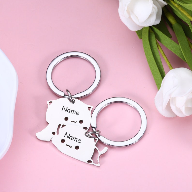 May include: Two silver keychains with a cat design. The keychains are connected and have the word "Name" engraved on them.