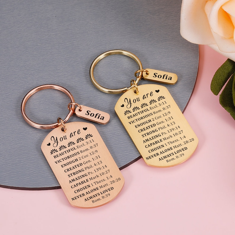 Puede incluir: Dos llaveros de tono dorado con dijes en forma de placa de identificaci&oacute;n. Los dijes tienen el texto "You are" seguido de una lista de afirmaciones positivas y vers&iacute;culos b&iacute;blicos. Los llaveros est&aacute;n personalizados con el nombre "Sofia" en cada etiqueta.