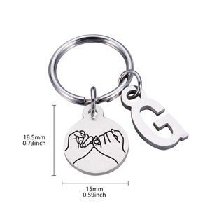 May include: Silver keychain with a circle charm featuring two hands making a pinky promise and a letter G charm. The keychain measures 0.73 inches in diameter and 0.59 inches in height.