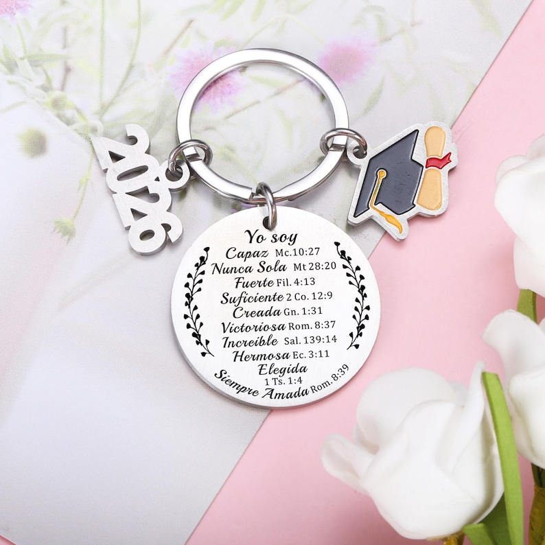 May include: A silver-toned metal keychain featuring a round pendant engraved with Spanish text and floral accents, a graduation cap charm, and a separate charm displaying the year 2026. The text includes phrases like Yo soy Capaz, Nunca Sola, and Fuerte.