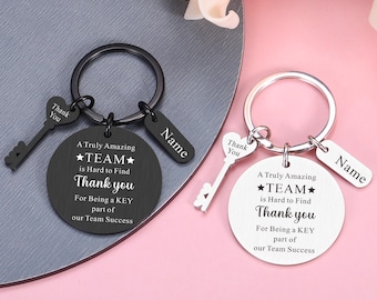 Könnte beinhalten: Zwei Metallschlüsselanhänger, einer in Schwarz und einer in Silber, jeweils mit einer runden Scheibe, die mit dem Text A Truly Amazing TEAM is Hard to Find Thank you For Being a KEY part of our Team Success, einem herzförmigen Schlüsselanhänger und einem Namensschild versehen ist.