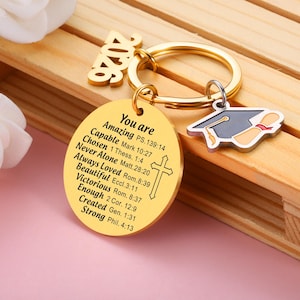 May include: A gold-toned metal keychain featuring a round disc engraved with the text You are Amazing PS.139:14, Capable Mark 10:27, Chosen 1 Thess. 1:4, Never Alone Matt.28:20, Always Loved Rom.8:39, Beautiful Eccl.3:11, Victorious Rom. 8:37, Enough 2 Cor. 12:9, Created Gen. 1:31, Strong Phil. 4:13, a cross, a 2026 charm, and a graduation cap charm.