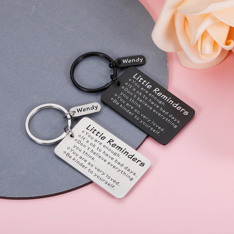 Op de afbeelding: Twee zilveren sleutelhangers met zwarte metalen labels. De labels hebben de tekst "Little Reminders" en inspirerende berichten. De sleutelhangers zijn gepersonaliseerd met de naam "Wendy".