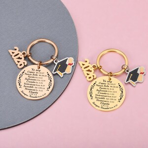 May include: Two gold-toned metal keychains featuring a circular pendant engraved with Spanish text and a leaf border. Each keychain includes a 2026 charm and a black and white graduation cap charm with a yellow tassel. The text reads Yo soy Capaz, Nunca Sola, Fuerte, Suficiente, Creada, Victoriosa, Incre&iacute;ble, Hermosa, Elegida, Siempre Amada.
