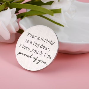 May include: A silver round token with the text "Your sobriety is a big deal, I love you & I'm proud of you."