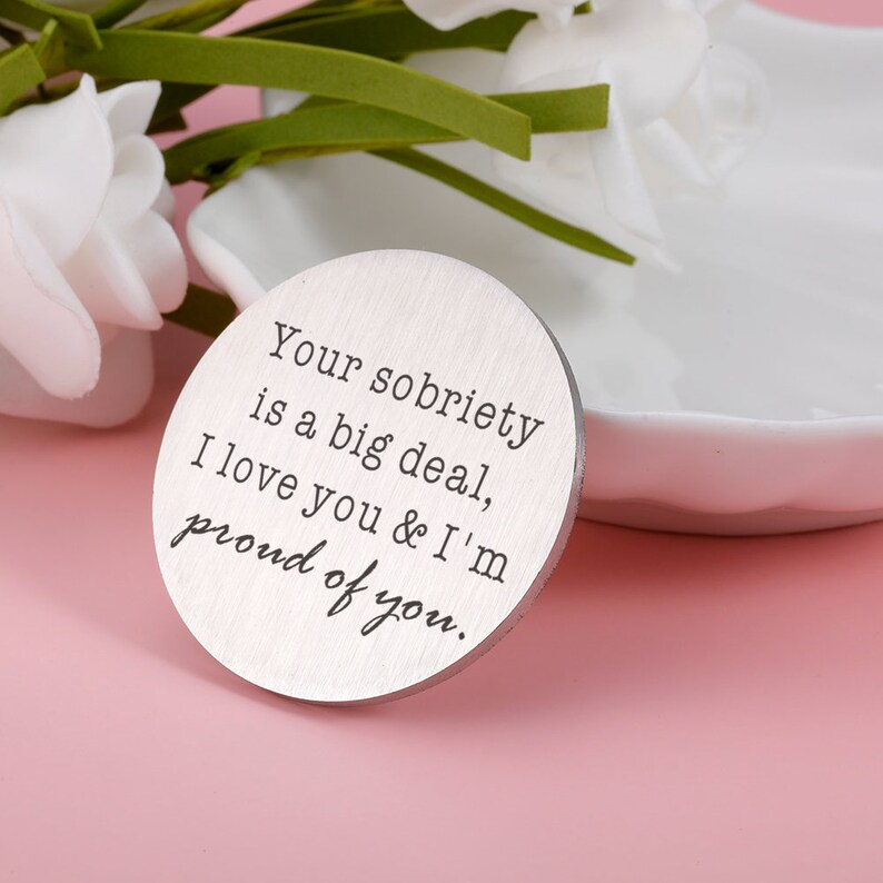 May include: A silver round token with the text "Your sobriety is a big deal, I love you & I'm proud of you."