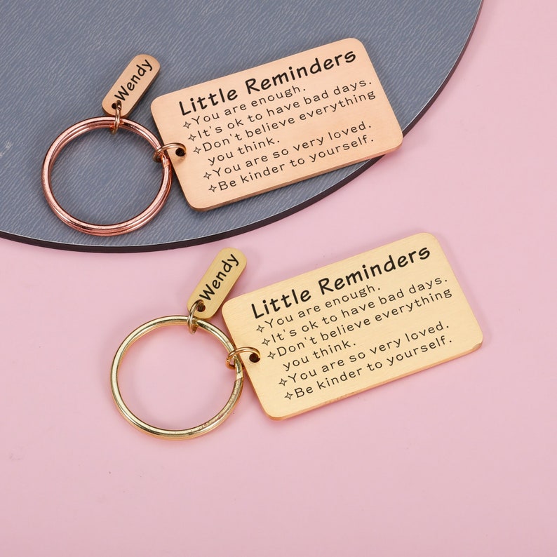 Op de afbeelding: Gouden sleutelhanger met een rechthoekig label waarop "Little Reminders" staat en positieve affirmaties bevat. Het label is bevestigd aan een gouden sleutelring met een kleiner label waarop "Wendy" staat.