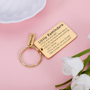 Op de afbeelding: Gouden sleutelhanger met een klein rechthoekig label met de tekst "Little Reminders" en een lijst met positieve affirmaties. Het label is bevestigd aan een gouden ring en een kleiner label met de naam "Wendy" erop.