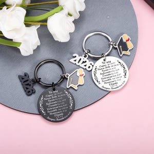 May include: Two circular metal keychains, one black and one silver, each featuring the number 2026, a graduation cap charm, and a round pendant engraved with the Spanish text Yo soy Capaz, Nunca Sola, Fuerte, Suficiente, Creada, Victoriosa, Incre&iacute;ble, Hermosa, Elegida, Siempre Amada.
