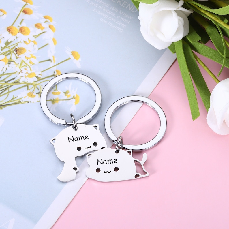 May include: Two silver keychains with cat-shaped charms. The charms have the word "Name" engraved on them. The keychains are on a pink and white background.