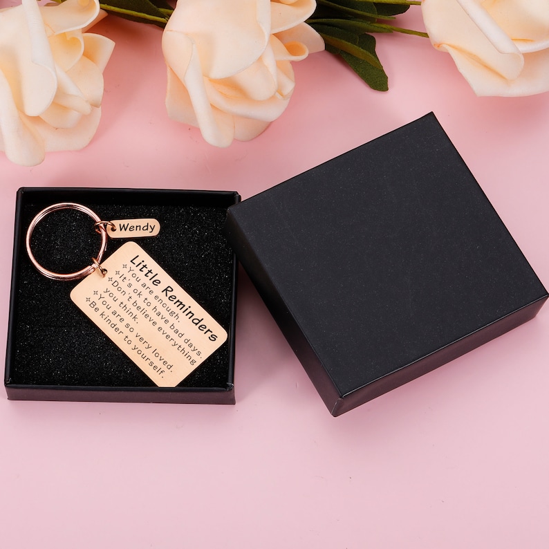 Op de afbeelding: Een gouden sleutelhanger met een zwarte doos. De sleutelhanger heeft een klein rechthoekig label met de tekst "Little Reminders" en de volgende tekst: "You are enough. It's ok to have bad days. Don't believe everything you think. You are so very loved. Be kinder to yourself."