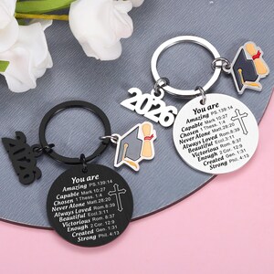 May include: Two circular metal keychains, one black and one silver, each featuring a graduation cap charm, the year 2026, and a cross. The discs are engraved with the text: You are Amazing PS.139:14, Capable Mark 10:27, Chosen 1 Thess. 1:4, Never Alone Matt.28:20.
