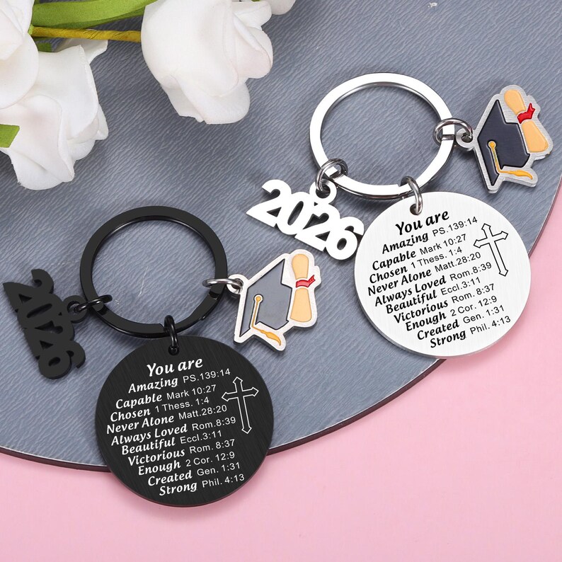 May include: Two circular metal keychains, one black and one silver, each featuring a graduation cap charm, the year 2026, and a cross. The discs are engraved with the text: You are Amazing PS.139:14, Capable Mark 10:27, Chosen 1 Thess. 1:4, Never Alone Matt.28:20.