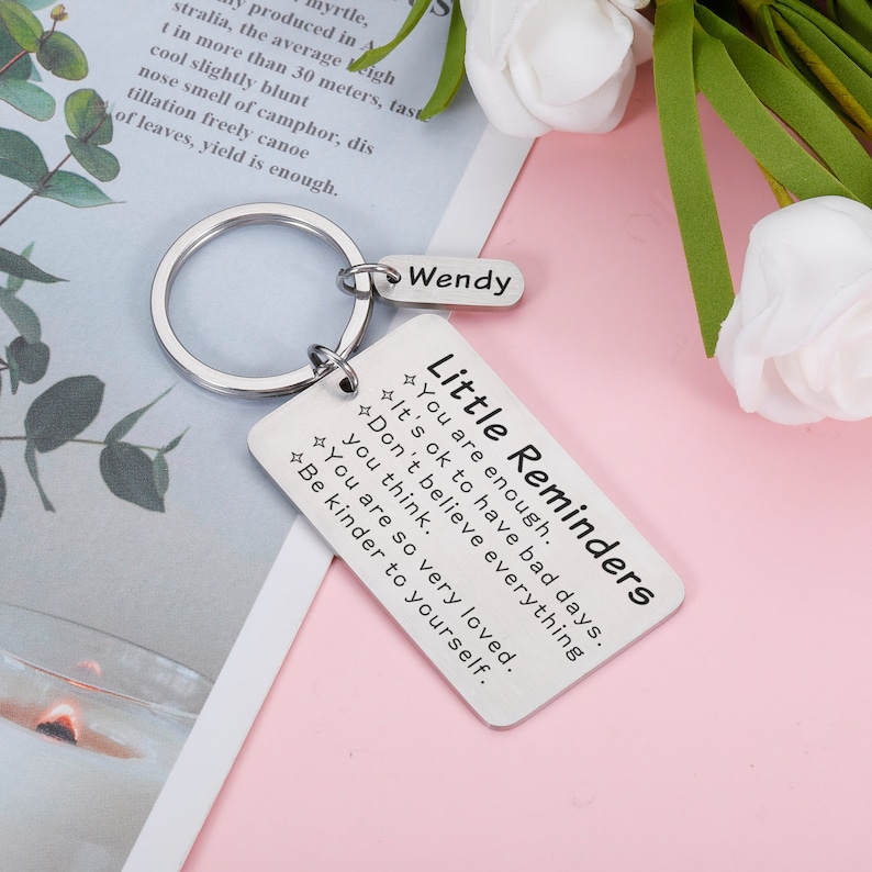 Op de afbeelding: Een zilveren sleutelhanger met een rechthoekig label waarop "Kleine herinneringen" staat en de tekst "Je bent genoeg. Het is ok&eacute; om slechte dagen te hebben. Geloof niet alles wat je denkt. Je bent zo geliefd. Wees vriendelijker tegen jezelf." De sleutelhanger heeft ook een kleiner label met de tekst "Wendy".