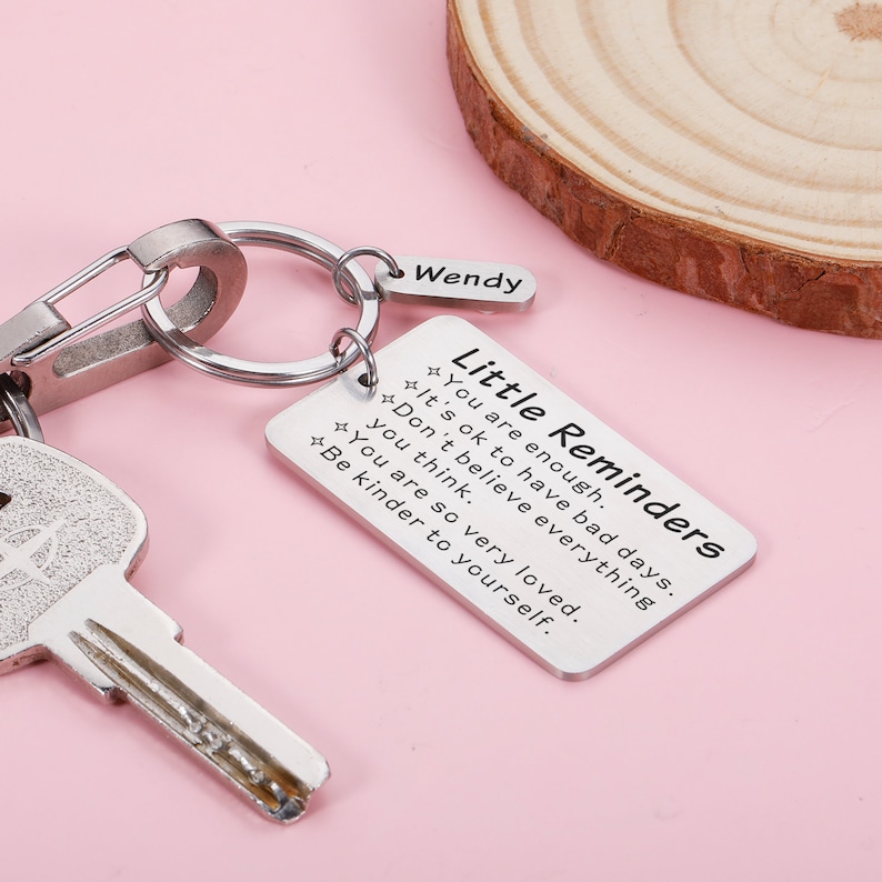 Op de afbeelding: Zilveren sleutelhanger met een klein rechthoekig label met de tekst "Kleine herinneringen" en de tekst "Je bent genoeg. Het is ok&eacute; om slechte dagen te hebben. Geloof niet alles wat je denkt. Je bent zo erg geliefd. Wees vriendelijker tegen jezelf." De sleutelhanger heeft ook een klein label met de tekst "Wendy".