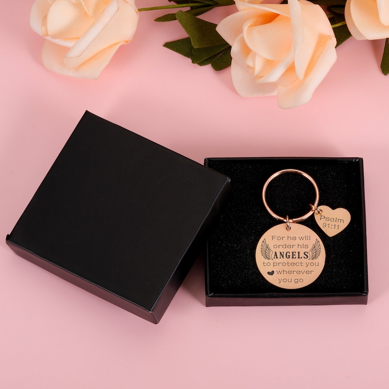 May include: A rose gold keychain with a round charm that reads "For he will order his angels to protect you wherever you go" and a heart-shaped charm that reads "Psalm 91:11". The keychain is in a black gift box.