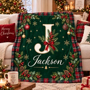 Könnte beinhalten: Eine dunkelgrüne Wohndecke mit einem großen cremefarbenen Buchstaben J, dem Namen Jackson in weißer Schreibschrift und einem festlichen Rand aus Tannenzweigen, roten Beeren, Tannenzapfen, goldenen Kugeln und Schneeflocken mit rot-grünem Karomuster.