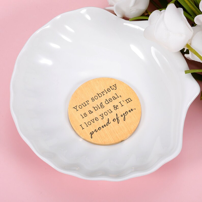 May include: A wooden circle with the text "Your sobriety is a big deal, I love you & I'm proud of you." The circle is sitting on a white seashell-shaped dish.