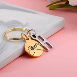 May include: Gold keychain with a round charm featuring two hands intertwined with a pinky promise. The charm is attached to a gold key ring and a silver letter H charm.