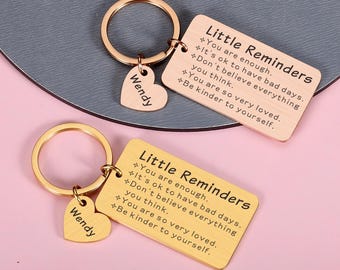 May include: Two rectangular metal keychains with round rings and heart-shaped charms engraved with the name Wendy. The rectangular tags feature the text Little Reminders followed by five positive affirmations. One keychain is rose gold and one is gold.