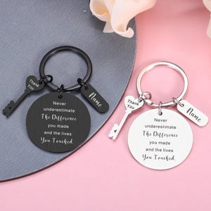May include: Two circular metal keychains, one black and one silver, each featuring a heart-shaped key charm engraved with Thank You, a rectangular tag with the word Name, and a round pendant inscribed with Never underestimate The Difference you made and the lives You Touched.