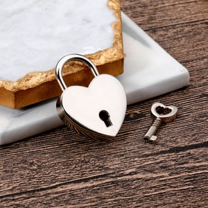 May include: A silver heart-shaped padlock with a keyhole and a matching silver key. The padlock and key are on a white marble surface.