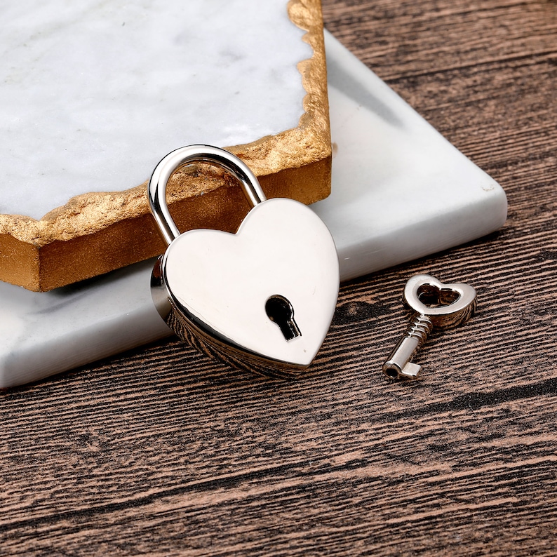 May include: A silver heart-shaped padlock with a keyhole and a matching silver key. The padlock and key are on a white marble surface.