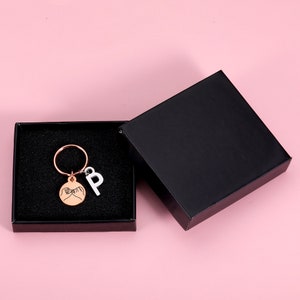 May include: A black gift box with a rose gold keychain inside. The keychain features a round charm with two hands making a pinky promise and a letter "P" charm.
