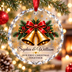 May include: A round, clear, faceted glass Christmas ornament featuring a red ribbon bow, green pine branches, red berries, and two gold bells. The text reads: Sophia & William, OUR FIRST CHRISTMAS TOGETHER, with a small gold heart.
