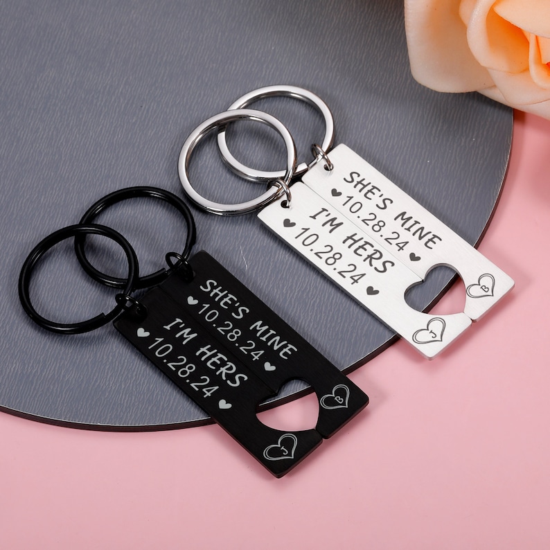 May include: Two black and silver keychains with interlocking heart shapes. The black keychain says "She's Mine 10.28.24" and "I'm Hers 10.28.24". The silver keychain says "She's Mine 10.28.24" and "I'm Hers 10.28.24".