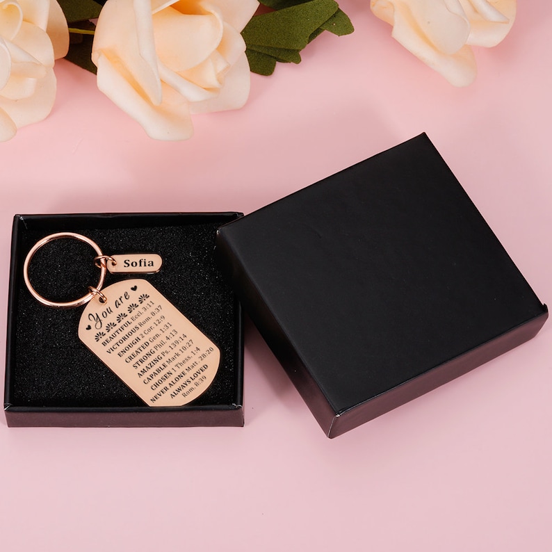 Puede incluir: Un llavero de oro rosa con una caja negra. El llavero tiene una peque&ntilde;a etiqueta con el nombre "Sofia" y una etiqueta m&aacute;s grande con el texto "You are" seguido de una lista de afirmaciones positivas y vers&iacute;culos b&iacute;blicos.