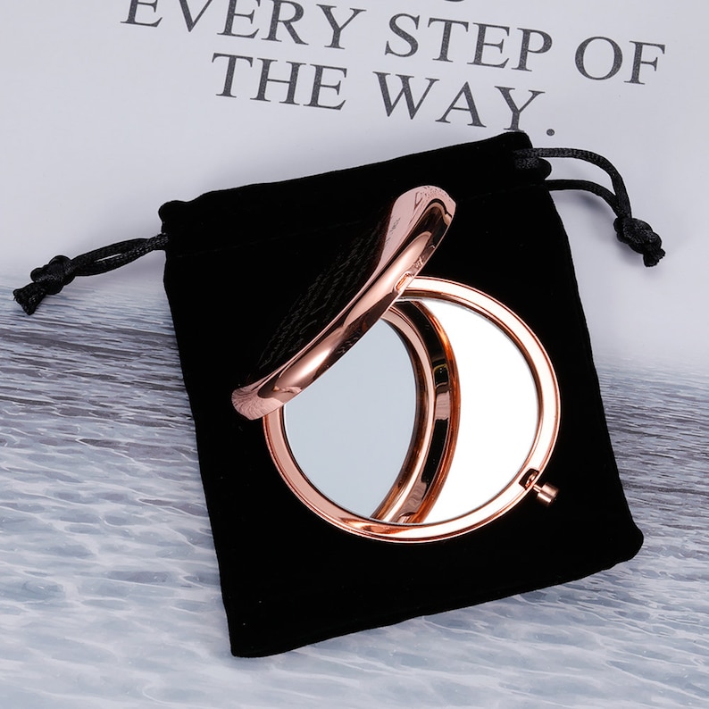 May include: A rose gold compact mirror with a black velvet pouch. The mirror is open and the reflective surface is visible.